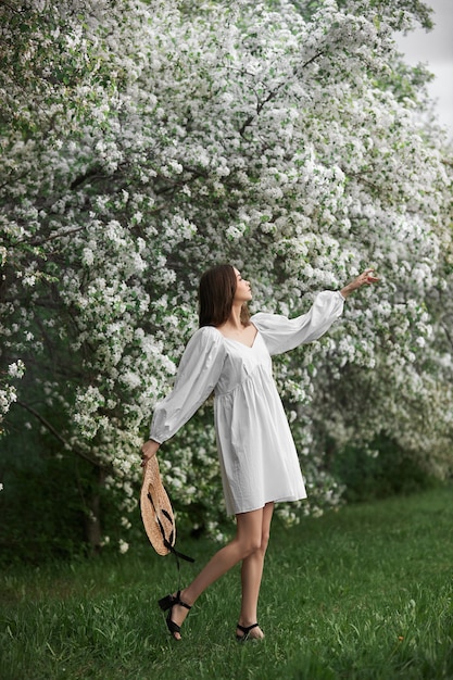 Молодая женщина в белом платье с соломенной шляпой гуляет по цветущему весеннему саду. Пришла весна, романтическое настроение