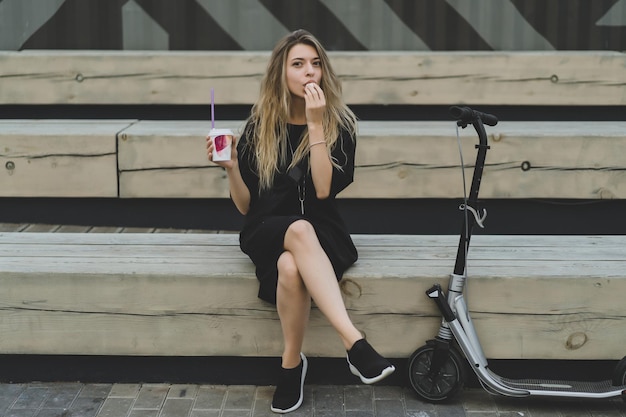 Молодая женщина с длинными волосами на электрический скутер. Девушка на электрическом самокате пьет кофе.