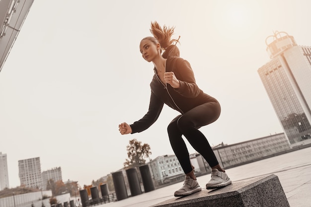 Фото Молодая женщина делает упражнения для ног на забитой камнями платформе шаг снаружи с городом на фоне. красивая женщина cauceuropean перепрыгивает через большой шаг, упражнение на корточки. концепции фитнеса и здоровья