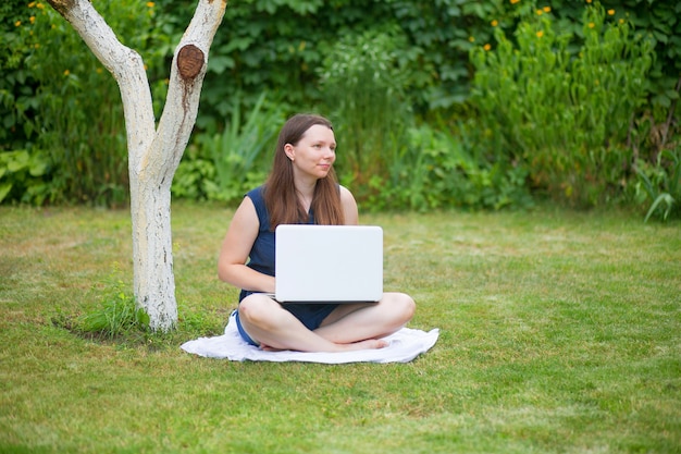 молодая женщина сидит на лугу с ноутбуком и разговаривает на онлайн-конференции на работе