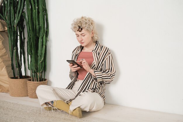 Young woman sitting on the floor near the exotic plants and typing a message online on her mobile phone