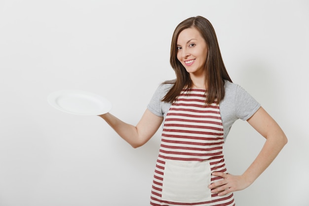 Домохозяйка молодого привлекательного усмехаясь брюнет кавказская в полосатом фартуке, серой изолированной футболке. Красивая домработница женщина держит белую пустую тарелку