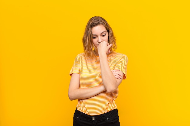 Photo young blonde woman feeling serious, thoughtful and concerned, staring sideways with hand pressed against chin against flat color wall