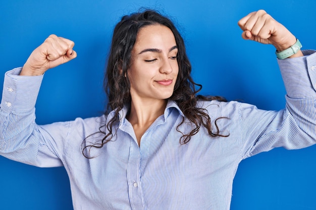 Молодая брюнетка женщина, стоящая на синем фоне, показывая мышцы рук, гордо улыбаясь фитнес-концепцией