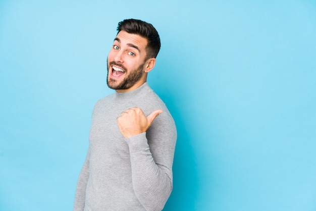 Young caucasian man against a blue wall isolated points with thumb finger away, laughing and carefree.