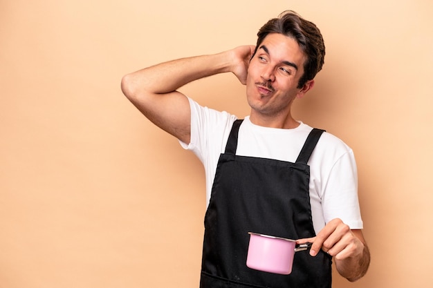 Young caucasian man holding a saucepan isolated on beige background touching back of head thinking and making a choice