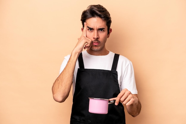 Young caucasian man holding a saucepan isolated on beige background pointing temple with finger, thinking, focused on a task.