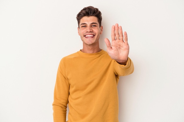 Молодой кавказский мужчина на белом фоне весело улыбается, показывая пальцами номер пять.