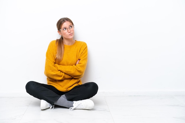 Photo young caucasian woman sitting on the floor isolated on white background making doubts gesture while lifting the shoulders