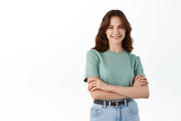 Фото Молодая уверенная в себе студентка в футболке скрестила руки на груди, улыбается и смотрит в камеру, стоящую на белом фоне