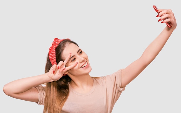 Фото Молодая европейка в повседневной одежде и красной повязке делает автопортрет над белой стеной студии счастливая женщина улыбается и делает селфи на своем смартфоне
