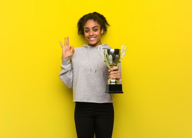 Фото Молодая чернокожая женщина фитнеса жизнерадостная и уверенно делая одобренный жест. держа трофей.