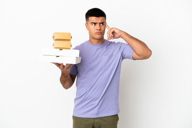 Photo young handsome man holding pizzas and burgers over isolated white background having doubts and thinking