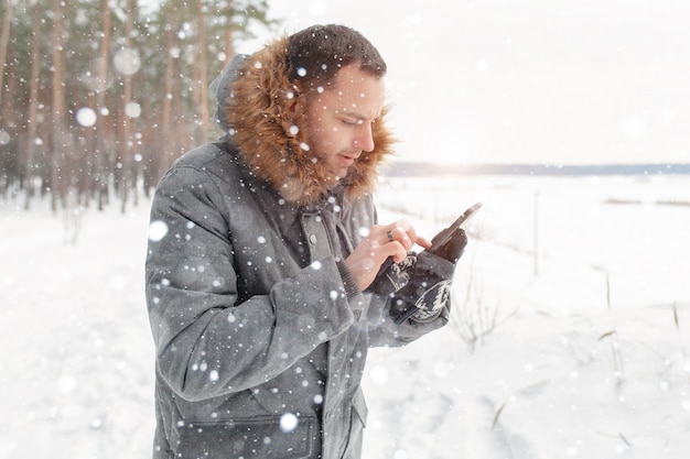 Молодой красавец использует мобильный телефон в снежном лесу.
