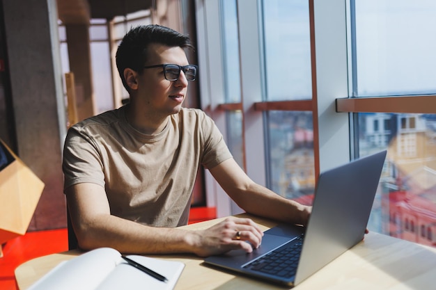 안경을 쓴 젊고 잘생긴 스마트 프리랜서는 카페에서 시간을 보내면서 노트북에서 일합니다 긍정적인 학생은 카페의 나무 테이블에 앉아 있습니다 실내에서 일하는 동안 앉아있는 행복한 남자