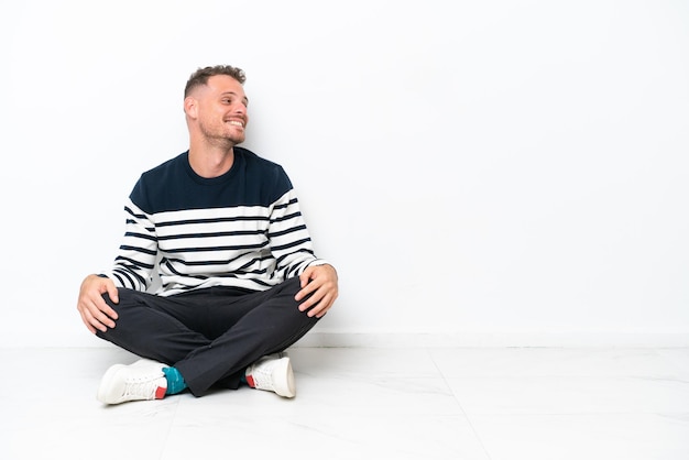 Photo young man sitting on the floor isolated on white background laughing in lateral position