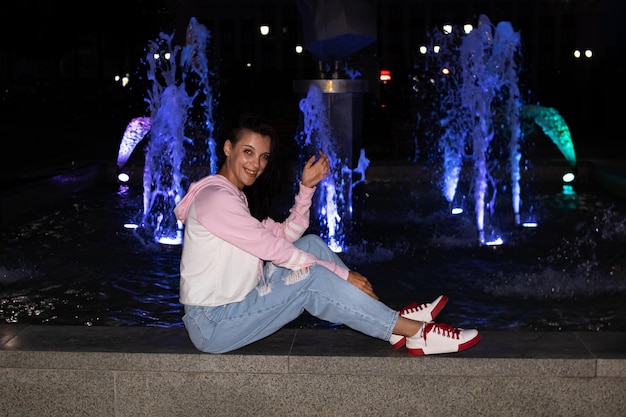 Молодая улыбающаяся девушка сидит ночью у фонтана