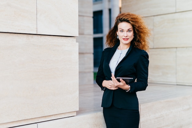 Молодая успешная деловая женщина в элегантной одежде, с пышными волосами, держа в руках планшет во время работы