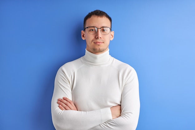 Foto zelfverzekerde man in bril poseren camera kijken met gevouwen armen, gekleed in wit casual shirt, geïsoleerd op blauwe studio achtergrond