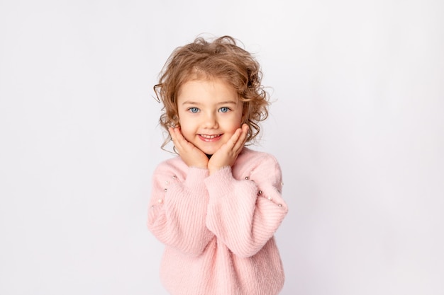 Фото Красивая детская девочка на белом изолированном фоне в розовом свитере сложила милые руки под щеками и улыбается, место для текста
