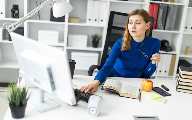 Фото Молодая девушка сидит за столом, держит в руке очки и работает за компьютером
