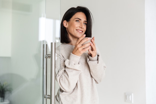 Afbeelding van volwassen vrouw 30s kopje met thee te houden, terwijl staande in de moderne lichte kamer in de ochtend