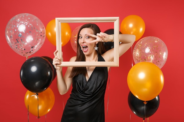 Пораженная молодая девушка в черном платье празднует показ знака победы, держащего рамку на красном фоне воздушных шаров. Международный женский день с новым годом, концепция вечеринки по случаю дня рождения макет.