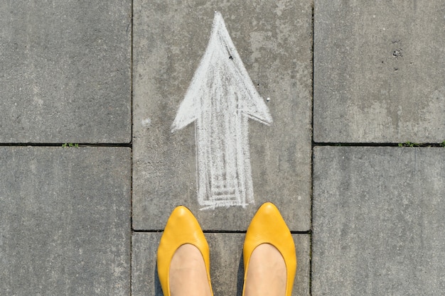 Знак стрелки нарисованный на сером тротуаре с женскими ногами