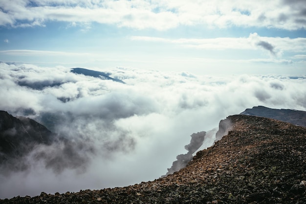 Atmospheric scenery on top of mountain ridge above clouds to vertex in thick low clouds. Minimalist view from precipice edge over clouds. Beautiful landscape with mountain range over dense clouds.
