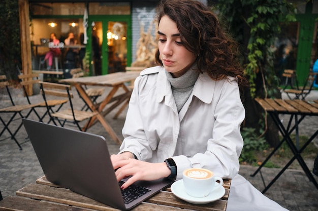 Привлекательная повседневная брюнетка в плаще уверенно учится на ноутбуке с кофе в кафе на открытом воздухе