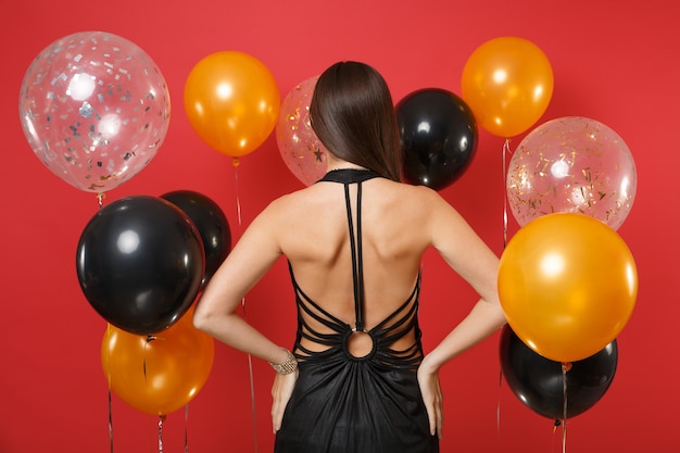 Вид сзади сзади молодой женщины в черном платье празднует, стоя с оружием подбоченясь на ярко-красном фоне воздушных шаров. День Святого Валентина, с новым годом, концепция вечеринки по случаю дня рождения макет.