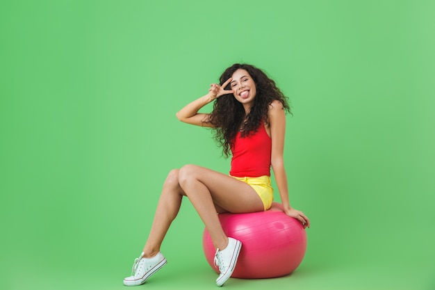 красивая женщина в летней одежде сидит на фитнес-мяче во время аэробики у зеленой стены
