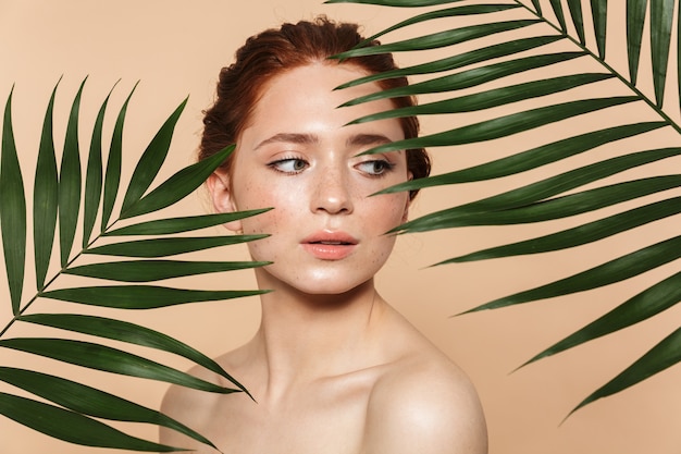 Photo beauty portrait of an attractive young topless redhead woman standing isolated, posing surrounded with tropical leaves