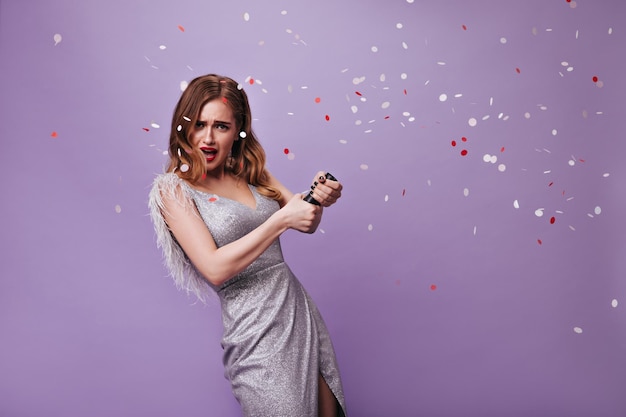 Блондинка в серебряном платье бросает конфетти Портрет кудрявой привлекательной женщины в праздничном наряде, смотрящей в камеру и веселящейся на фиолетовом фоне
