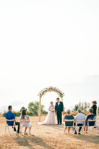 사진 신부 와 신랑 은 손님 들 앞 에 있는 등록관 근처 의 잔디 ⁇  에 있는 결혼 아치 에 서 있다