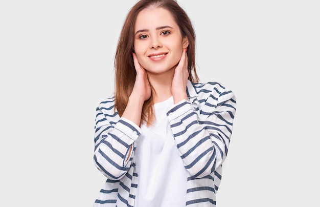 Откровенный снимок молодой женщины брюнте, позитивно улыбающейся в повседневной одежде и смотрящей в камеру с рукой на лице, изолированной на белой стене студии Эмоции людей