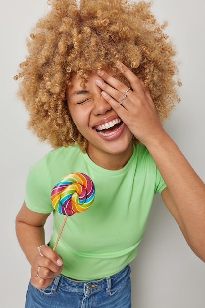 Photo carefree overjoyed millennial girl with curly hair makes face palm laughs gladfully holds multicolored round lollipop on stick dressed in casual green t shirt and jeans isolated over white wall