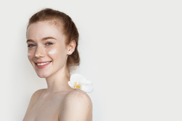 Photo caucasian woman with red hair and freckles is posing with a cream on her face at camera