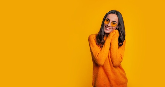 Очаровательная молодая женщина в желтом свитере и солнцезащитных очках позирует с улыбкой на желтом фоне и смотрит в камеру