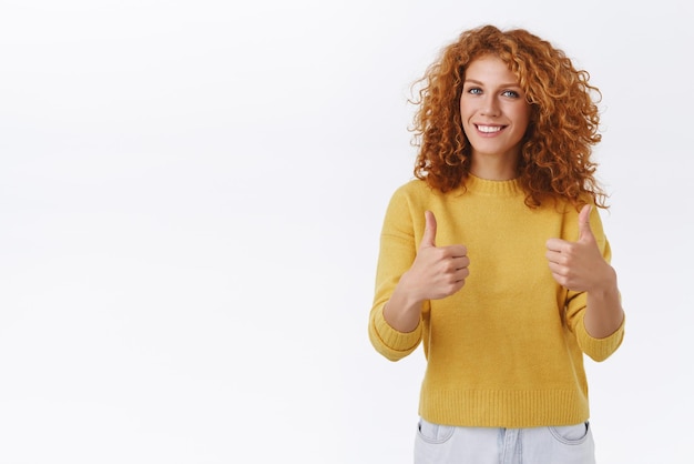 Веселая привлекательная кудрявая рыжая женщина в желтом свитере показывает большой палец в одобрении, подбадривая или поддерживая вас, оставляя положительный отзыв, рекомендует продукт, согласен, как событие на белом фоне