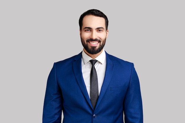 Photo cheerful bearded man with charming smile, standing and looking at camera, expressing positive emotions, wearing official style suit. indoor studio shot isolated on gray background.