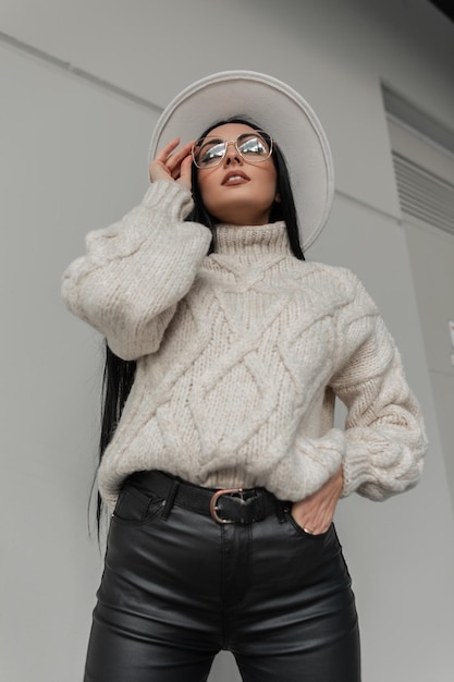 Фото Крутой городской портрет красивой девушки-модели в белом винтажном свитере, кожаных штанах и шляпе, надевающей очки возле серой стены на улице