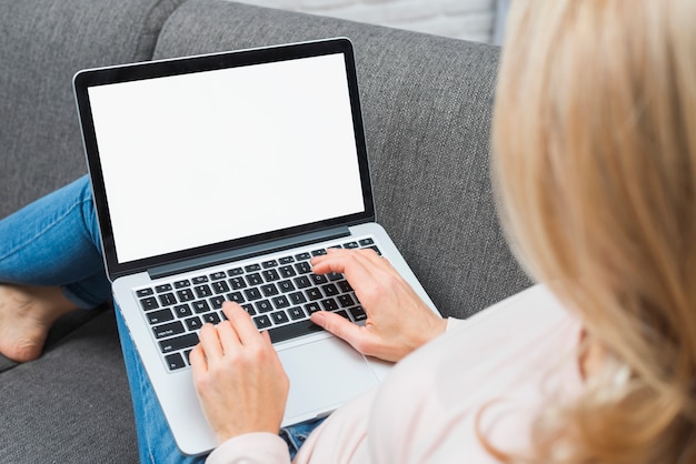 Close-up of blonde woman sitting on sofa using laptop with blank white screen