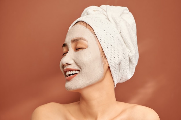 Photo close-up portrait of attractive girl with a towel on head and clay mask on face isolated over orange background.