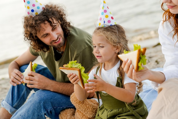 Крупным планом семья в шляпах для вечеринок ест бутерброды на пляже