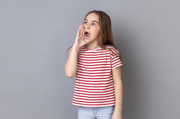 Темноволосая очаровательная маленькая девочка в полосатой футболке кричит громкую рекламу