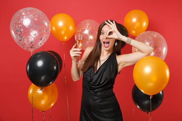 Фото Возбужденная девушка в черном платье празднует проведение бокала шампанского, показывая знак ок возле глаз на ярко-красном фоне воздушных шаров. женский день, с новым годом, концепция праздничной вечеринки макет дня рождения.