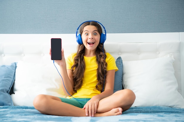 Выразительная эмоциональная взволнованная девочка-подросток показывает экран телефона Девочка-подросток в наушниках