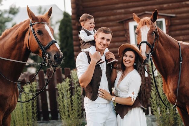 Семья в белых одеждах с сыном стоят возле двух красивых лошадей на природе. Стильная пара с ребенком фотографируются с лошадьми.