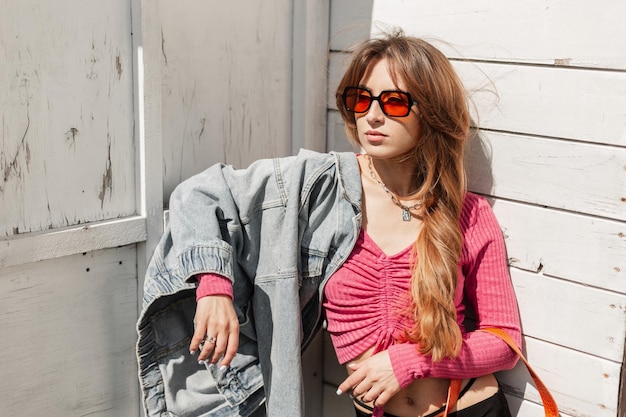 Photo fashionable beautiful model woman with fashion cool orange sunglasses in a stylish knit pink top with jeans jacket stands near a vintage white wooden building on the beach